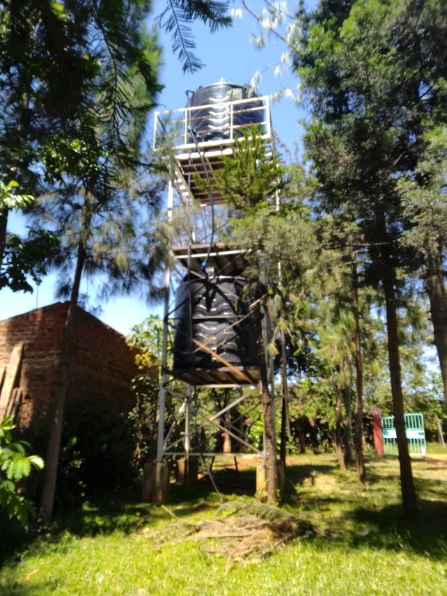 Solar-powered elevated water storage tanks among trees at a Shiraha Fresh Water distribution point